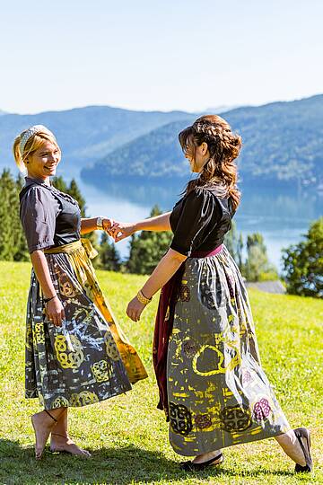 2 Frauen tanzen auf einer Wiese in Tracht mit Blick auf den Millstätter See
